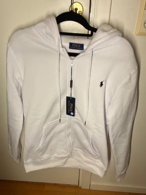 Vit hoodie från polo Ralph Lauren  - Vit hoodie från Polo Ralph Lauren med dragkedja, huva och snörning. Klassisk roterad logga på bröstet och känguruficka framtill. Tillverkad i ett mjukt bomullsmaterial, perfekt för en clean och stilren look.