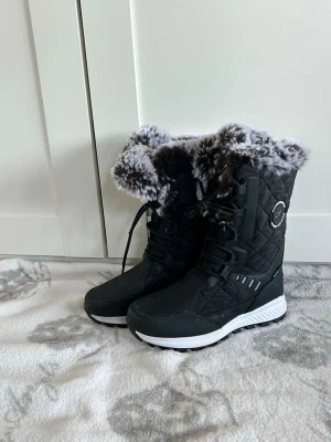 Svarta vinterstövlar med päls - Säljer ett par svarta vinterstövlar från Dawn Outdoor med quiltad design och snörning framtill. Insidan och skaftet är fodrade med mjuk, gråmelerad fuskpäls. Stövlarna har en vit platt sula med grovt mönster för extra grepp. Perfekta för kalla dagar.