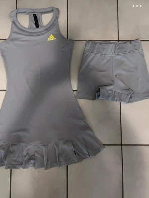 Grå Adidas courtdress padelklänning strl xs - Adidas courtdress  med matchande shorts. Klänningen har volangkant nedtill och ett gult Adidas-logo på bröstet. Perfekt för tennis eller träning, tillverkad i mjukt och stretchigt syntetmaterial för maximal komfort. XS