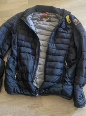 Svart quiltad dunjacka Parajumpers - Svart dunjacka från Parajumpers med quiltad design och coolt märke på axeln. Jackan har dragkedja framtill, två sidofickor med dragkedja och ljusgrått foder. Perfekt för kalla dagar och har en snygg, sportig look.Jackan har även två lufthål under armhålan och det finns i alla parajumper jackor.Jackan har ett problem och det är att kedjan saknar en sak men annars får funkar jacka extremt bra.