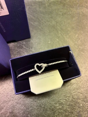 Swarovski silverarmband med hjärta - Snyggt armband från Swarovski i silverfärg med glittrande stenar och ett stort hjärta i mitten. Helt ovanvänt och orört. Nypris 1750kr