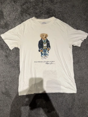 Vit Polo Bear t-shirt från Ralph Lauren - Vit t-shirt från Polo Ralph Lauren med ikoniskt Polo Bear-tryck på framsidan. Klassisk rund hals och korta ärmar. Mjuk bomullskvalitet som känns skön mot huden. Perfekt för dig som gillar stilrena och coola prints. Använd fåtal gånger