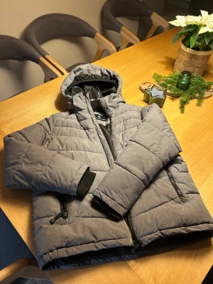 Grå pufferjacka från Cross Sportswear - Snygg grå pufferjacka från Cross Sportswear med huva och svarta detaljer. Jackan har dragkedja framtill, två fickor med dragkedja och mjukt foder vid halsen. Perfekt för kalla dagar och enkel att matcha med det mesta.