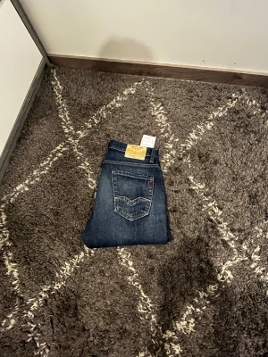 Replay blå jeans, modell 14A - Snygga blå jeans från Replay med klassisk femficksdesign och guldfärgade detaljer. Jeansen har en rak passform och tydliga Replay-loggor på bakficka och läderpatch. Perfekta för dig som gillar stilrena och tidlösa jeans.