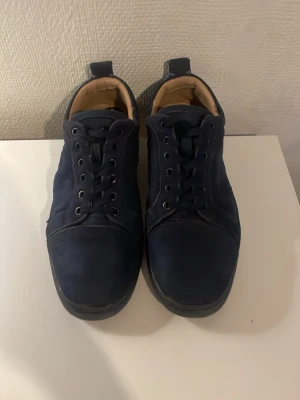 Mörkblå sneakers från Louboutin - Säljer ett par mörkblå sneakers från Christian Louboutin med klassisk röd sula och broderad logga på hälen. Skorna har snörning, rund tå och detaljer i skinn samt guldiga öljetter på sidan. Insidan är beige och materialet känns lyxigt och mjukt.