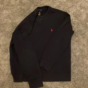 Mörk blå sweatshirt från Polo Ralph Lauren - Mörk blå sweatshirt från Polo Ralph Lauren med klassisk röd broderad logga på bröstet. Tröjan har rund halsringning, långa ärmar och ribbade muddar. Perfekt för en stilren och avslappnad look.