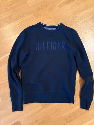 Marinblå Tommy Hilfiger sweatshirt S - Snygg marinblå sweatshirt från Tommy Hilfiger i vintage fit. Tröjan har ett stilrent HILFIGER-tryck på bröstet och klassisk rund hals med diskreta ränder på insidan. Mjukt material och långa ärmar, perfekt för chill dagar.