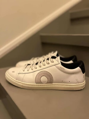 Oliver Cabell Low 1 Belmont sneakers - Sparsamt använda! Snygga vita Oliver Cabell Low 1 Belmont sneakers i skinn med grå mockadetalj i form av en cirkel på sidan. Platt sula, rund tå och svart häl med guldfärgad logga. Klassisk siluett med snörning och stilren design, perfekt för dig som gillar minimalistiska sneakers.