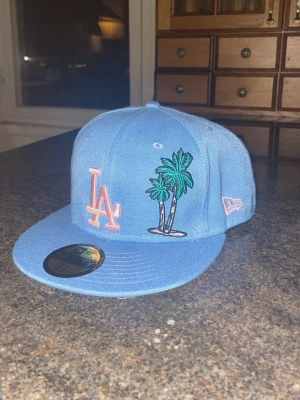 Ljusblå keps - Bilderna visar en blå Los Angeles Dodgers keps från New Era, modell 59FIFTY, med broderade palmer och jubileumsmärke. Ingen väst syns på bilderna.