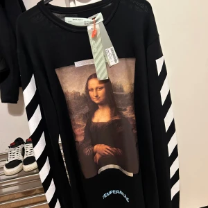 Off-White Mona Lisa - Säljer en svart långärmad tröja från Off-White med ikoniskt Mona Lisa-print på framsidan och stora vita pilar på ryggen. Ärmarna har vita diagonala ränder och texten 'TEMPERATURE' i blått nedtill.