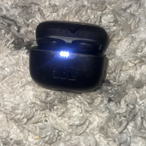 JBL trådlösa hörlurar med laddningsetui - Snygga svarta JBL trådlösa hörlurar med laddningsetui. Laddningsindikatorn lyser och etuiet har USB-C port. Viss kosmetisk slitage syns på etuiet men fungerar utmärkt. Perfekt för musik och samtal på språng.