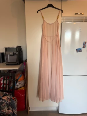 Ljusrosa långklänning från Ida Sjöstedt - Drömmig ljusrosa långklänning från Ida Sjöstedt med tunna axelband dekorerade med guldfärgade detaljer. Klänningen har ett flytande, skirt tyg i polyester och ett smalt bälte med guldfärgad spänne i midjan. Perfekt för dig som gillar romantisk och elegant stil. Storlek M. Mycket fint skick, jag hittar inga synliga anmärkningar eller defekter. 