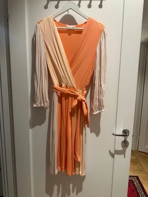 Omlottklänning från Calvin Klein stl XS - Snygg omlottklänning från Calvin Klein i ljus orange och beige med v-ringning och knytband i midjan. Klänningen har trekvartsärmar med lätt puff och är tillverkad i mjukt, följsamt material som ger ett skönt fall. Perfekt för dig som gillar färg och stil.