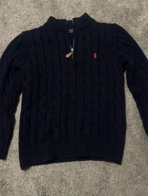 Polo Ralph Lauren quarter zip - Kabelstickad mörkblå tröja från Polo Ralph Lauren med halv dragkedja och röd broderad logga på bröstet. Tröjan har ribbade muddar, hög krage och är långärmad. Perfekt för kyliga dagar och snygg till jeans. Är L men passar mer som en M då den är tvättad en del. Hör av er vid minsta lilla fråga och priset är ej hugget i sten