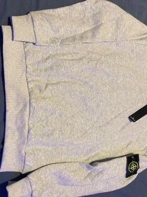 Grå sweatshirt från Stone Island - Snygg ljusgrå sweatshirt från Stone Island med klassisk kompasspatch på ärmen. Tröjan har rund hals, ribbade muddar och är tillverkad i mjuk bomull. Perfekt för dig som vill ha en clean och stilren look med streetkänsla.