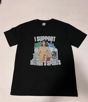 ”I Support Women's Sports” T-shirt  - En svart t-shirt med ett humoristiskt motiv. Trycket föreställer en kvinna framför en tvättmaskin tillsammans med texten “I Support Women’s Sports” i stora, tydliga bokstäver. Perfekt för dig som gillar satiriska tryck. Tröjan är i nytt skick och sitter bekvämt.