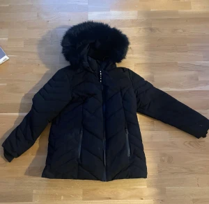 Svart pufferjacka med fuskpäls från Amisu - Snygg svart pufferjacka från Amisu med en stor huva och fluffig fuskpäls. Jackan har dragkedja framtill, två fickor med dragkedja och ett quiltat mönster. Perfekt för kalla vinterdagar och riktigt mysig att bära.