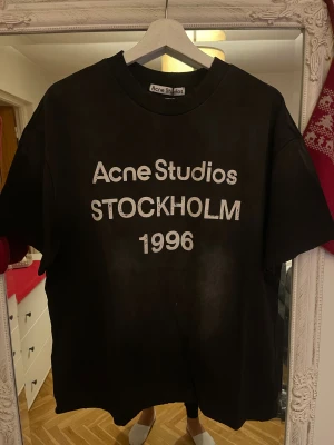 Svart Acne Studios t-shirt Stockholm - Cool svart t-shirt från Acne Studios med vit text 'Acne Studios STOCKHOLM 1996' på bröstet. Oversized fit och klassisk rund hals. Perfekt statement-plagg för dig som gillar streetstyle och skandinavisk design.