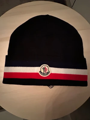 Moncler mössa med röd, vit och blå rand - Snygg Moncler mössa i mörkblått med breda ränder i rött, vitt och marinblått längs kanten. Framtill sitter Moncler-loggan broderad i vitt med röd och blå detalj. Mössan är stickad och har en klassisk, rundad form som passar perfekt till vinterlooken.
