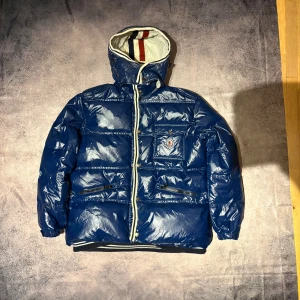 Blå Moncler pufferjacka med huva - Säljer en blå pufferjacka från Moncler med glansig finish, huva med röd och marinblå rand på insidan, vita detaljer längs dragkedjan och flera fickor framtill. Jackan har Moncler-logga på bröstfickan och ribbad mudd vid nederkant.