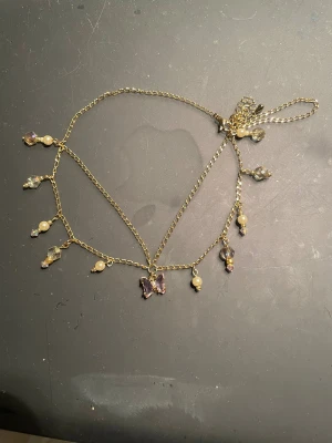 Guldig halskedja med fjäril och pärlor - Guldig kedja med hängande pärlor och glittriga stenar, samt en lila fjäril i mitten. 