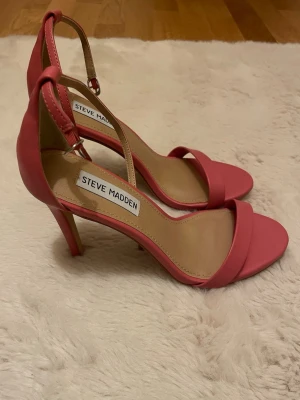 Steve Madden rosa klackpumps - Snygga rosa pumps från Steve Madden med smal rem runt ankeln och öppen tå. Skorna har en elegant hög klack och är tillverkade i slätt syntetmaterial med beige innersula. Perfekt för dig som gillar stilrena och trendiga skor med feminin touch.