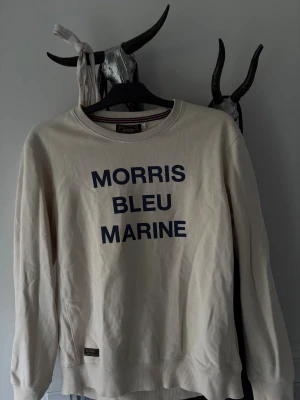 Beige sweatshirt från Morris - Snygg beige sweatshirt från Morris med mörkblå text 'MORRIS BLEU MARINE' på bröstet. Tröjan har rund hals, långa ärmar och ribbade muddar. Perfekt för dig som gillar stilrena och klassiska plagg med en twist.