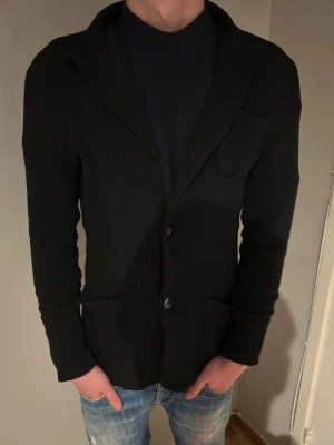John Henric Svart stickad kavaj med fickor - Svart stickad kavaj med två stora framfickor och klassisk krage. Kavajen har knäppning med två knappar och en enkel, clean look som passar till jeans eller chinos. Perfekt för dig som vill ha en avslappnad men ändå stilren vibe.