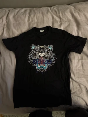 Svart Kenzo t-shirt med tigertryck - Svart t-shirt från Kenzo med ikoniskt tigertryck i vitt, blått och turkost på bröstet. Klassisk rund hals och korta ärmar. Materialet är mjuk bomull och trycket har coola detaljer som gör att den sticker ut.