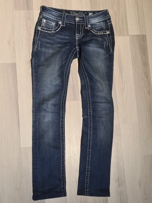 Miss Me blå jeans med strassdetaljer stl 24 - Snygga blå jeans modell straight från Miss Me i stl 24 med coola strassdetaljer och broderier på fickorna. Jeansen har kontrastsömmar, låg midja och klassisk femficksdesign. Materialet är stretchigt denim för skön passform. Perfekt för dig som gillar att sticka ut med detaljer.