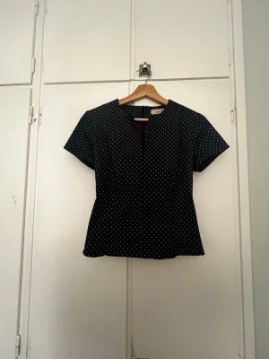 Svart prickig topp med v-ringning - Svart blus med vita prickar och v-ringad hals. Kortärmad och lätt figursydd modell med rå kant nedtill. Materialet känns som bomull eller linne. Snygg att matcha med vad som, jeans till exempel. Köpt på loppis men aldrig använd. Ser ut som ny förutom att jag har klippt av den längst ner. Då det var en klänning tidigare.