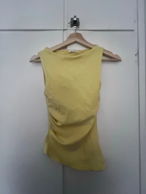 Gul figursydd topp Zara - Det var lite svårt att visa färgen på kameran men den är en ljusgul nyans, ljusare än på bilderna.                                                         Snygg ljusgul linnetopp från Zara med asymmetrisk drapering på ena sidan. Toppen är ärmlös och har en rund halsringning. Materialet är mjukt och lätt, vilket gör den skön att bära den. Passar vardagligt och även lite uppstylat med kostymbyxor exempletvis på finare tillfällen. Skit snygg. Köp gärna.