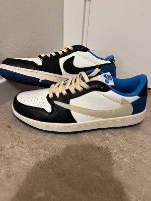 Nike Air Jordan 1 Low Travis Scott - Nike Air Jordan 1 Low Travis Scott i svart, vit och blå med beige snörning. Skorna har omvänd Swoosh på sidan, broderad Cactus Jack-logga på hälen och Air Jordan Wings på andra hälen. Platt blå sula och rund tå. Tillverkade i skinn med blå textilfoder.