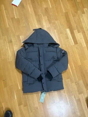 Grå black label canada goose macmillan L - Säljer en mörkgrå dunjacka från Canada Goose med huva och klassisk patch på ärmen. Jackan har dragkedja och knappar framtill, stora fickor och ribbade muddar vid ärmsluten. Perfekt för kalla dagar och riktigt snygg streetstil. Pris är ej hugget i sten!