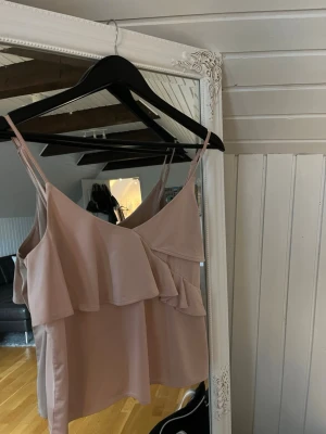 Ljusrosa linnetopp med volang från H&M - Supersöt ljusrosa linnetopp från H&M med dubbla smala axelband och volangdetaljer framtill. Toppen har en v-ringad hals och är tillverkad i mjuk polyester som känns skön mot huden. Perfekt för sommarens alla tillfällen!