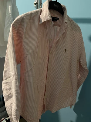 Rosa och vit randig skjorta Ralph Lauren - Klassisk skjorta från Ralph Lauren Sport i rosa och vitt med smala ränder. Skjortan har lång ärm, knappar framtill och ikonisk broderad logga på bröstet. Tillverkad i mjuk bomull och har en tidlös krage.