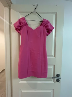 Rosa kort klänning med puffärmar - Superfin rosa kort klänning med markerad midja och stora puffärmar. Klänningen har en v-ringad urringning och dragkedja i ryggen. Materialet är slätt och glansigt på insidan, troligtvis polyester. Perfekt för dig som vill sticka ut med en färgstark look. Behöver bara att strykas lite. Har bara använt en gång till en fest!