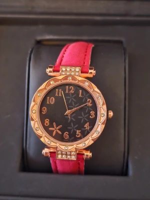 Röd klocka med blommönster och strass - Snygg klocka med rött band och roséguldfärgat ur med strassdetaljer runt boetten. Svart urtavla med blommönster och guldiga siffror. Bandet är i syntetmaterial och har klassiskt spänne i silver. Perfekt accessoar för att lyfta din stil.