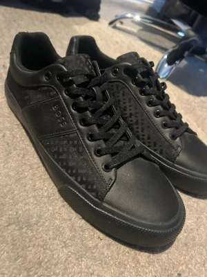 Svarta sneakers från BOSS - Snygga svarta sneakers från BOSS med diskret mönster på sidorna och tungan. Skorna har rund tå, platt sula och snörning framtill. Materialet är en mix av skinn och syntet, vilket ger en cool och modern look. Perfekta för dig som gillar stilrena och trendiga skor.