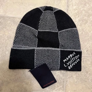 Grå och svart rutig Louis Vuitton väst - Snygg väst från Louis Vuitton i grått och svart med rutigt mönster. Västen har en modern design med kontrasterande paneler och ett broderat Louis Vuitton-tryck på framsidan. Perfekt för dig som gillar exklusiva märken och stilrena detaljer.