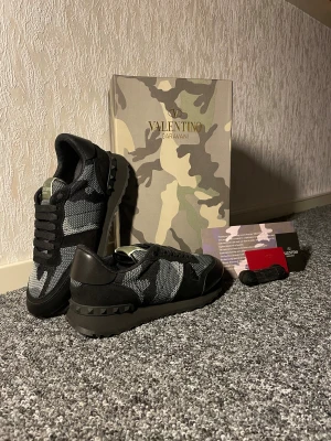 Valentino Garavani Rockrunner sneakers - Snygga Valentino Garavani Rockrunner sneakers i svart och grått med camouflagemönster. Skorna har platt sula, rund tå och coola nitar på hälen. Materialmix av mocka, skinn och textil. Kommer med originalkartong, extra skosnören och tags. Perfekt för dig som gillar exklusiv streetstyle.