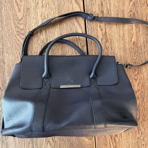 Axelremsväska - Större axelremsväska i mörkblått. Den har en stadig botten som är ca 16cm djup. Storleken är ungefär 40x25x16 cm. Färgen är mest rättvis på sista bilden. Den är i använt skick, men den har inga större defekter.