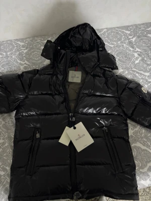 Svart Moncler pufferjacka med huva - Svart pufferjacka från Moncler med glansig finish och huva. Bilderna är tagna för 2 veckor sedan och prislappen är borttagen men jackan är helt oanvänd fortfarande. Passar dig som har XS i storlek och S framförallt. PRIS KAN DISKUTERAS
