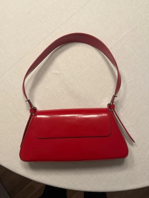 Röd axelväska  - Snygg röd axelväska med stilren, rektangulär form och bred axelrem. Använd en gång så i princip som ny.