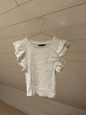 Vit ribbad topp med volangärmar - Säljer en vit ribbad topp från Zara med korta ärmar och snygga volangdetaljer på axlarna. Toppen har rund halsringning och är gjord i mjuk bomull. Perfekt för dig som gillar en clean look med lite extra edge på ärmarna.