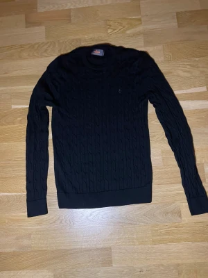 Svart kabelstickad tröja Morris  - Svart kabelstickad tröja från Morris i 100% merinoull. Klassisk rund halsringning, diskret broderad logga på bröstet och långa ärmar. Perfekt för dig som gillar stilrena och tidlösa plagg med premiumkänsla.