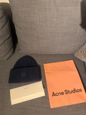 Mörkblå mössa från Acne Studios - Sprillans ny snygg mörkblå ribbstickad mössa från Acne Studios med klassisk logopatch framtill. Mössan är tillverkad i mjuk ull och har en uppvikt kant för extra stilpoäng. Perfekt accessoar för att hålla värmen och se cool ut under kyliga dagar.