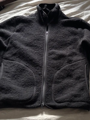 Svart fleecejacka  J.Lindeberg strl M - Svart fleecejacka  från J.Lindeberg med hög krage och hel dragkedja framtill. Materialet är mjukt och värmande fleece, perfekt för kyliga dagar. Snygga detaljer med J.Lindeberg-logga på ryggen och dragkedjan. Två stora fickor framtill för extra funktion.