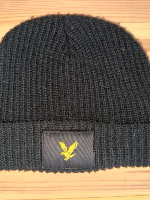 Svart stickad mössa från Lyle & Scott jättevarm - Snygg svart stickad mössa från Lyle & Scott med klassisk ribbad struktur. Framtill finns en svart patch med den ikoniska gula fågeln. Perfekt för att hålla värmen och addera stil till din outfit.
