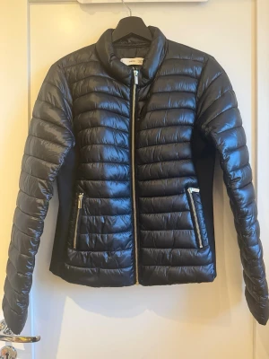 Svart pufferjacka från Mango - Snygg svart pufferjacka från Mangoi storlek M. Jackan har quiltad design, dragkedja framtill och två fickor med dragkedja. Perfekt för kyliga dagar och ger en clean, modern look. Materialet är glansigt och känns lätt men ändå värmande.
